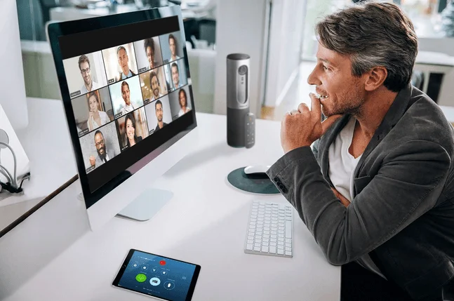 Man in an office on a Zoom video call, with a grid of participants on his desktop screen